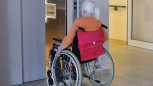 Ältere Frau im Rollstuhl in einem Innenbereich vor einem Aufzug.