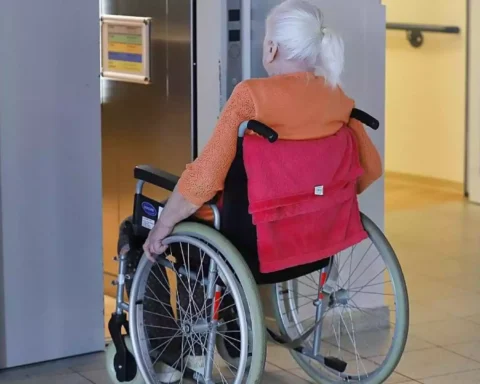 Ältere Frau im Rollstuhl in einem Innenbereich vor einem Aufzug.