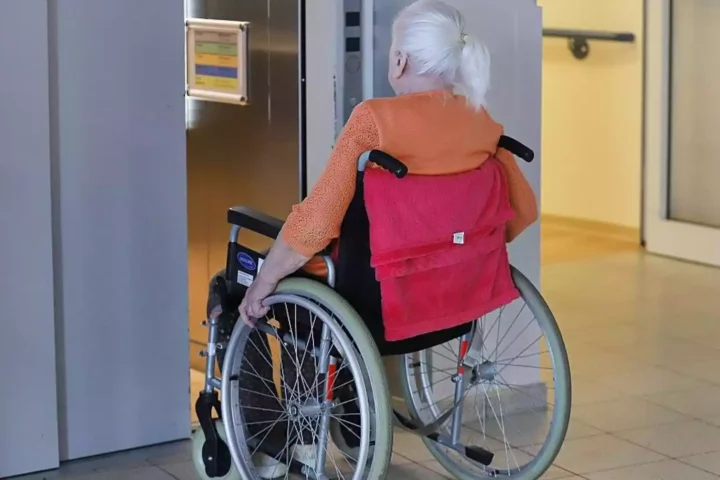 Ältere Frau im Rollstuhl in einem Innenbereich vor einem Aufzug.