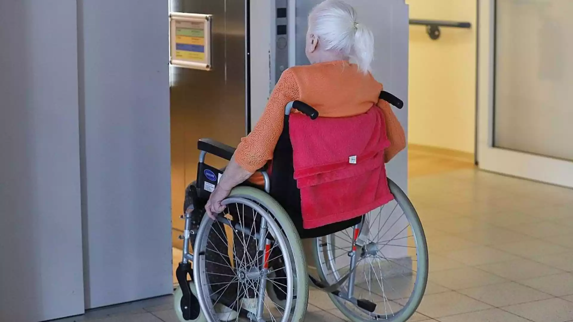 Forsa-Umfrage: Mehrheit lehnt höheren Kinderlosenzuschlag in der Pflege ab 1 Ältere Frau im Rollstuhl in einem Innenbereich vor einem Aufzug.