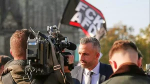 Sahra Wagenknecht spricht vor Kameraleuten bei einer politischen Veranstaltung, im Hintergrund eine Fahne mit „FCV AFD“.