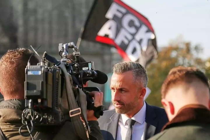 Sahra Wagenknecht spricht vor Kameraleuten bei einer politischen Veranstaltung, im Hintergrund eine Fahne mit „FCV AFD“.