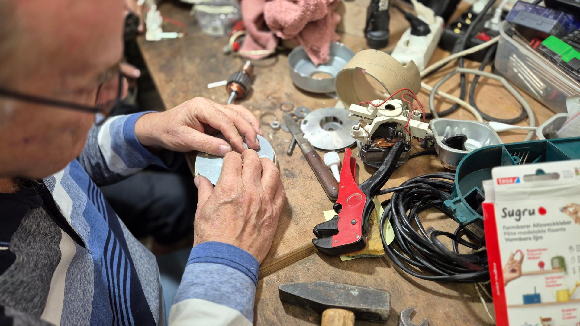 Ein Mann repariert an einem Arbeitstisch ein elektronisches Gerät mit Werkzeugen und Kabeln.