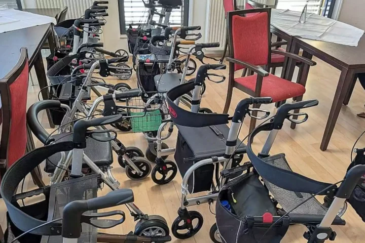 Mehrere Rollatoren stehen in einem hellen Raum zwischen Stühlen und Tischen.