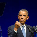 Barack Obama spricht an einem Podium mit Mikrofonen vor blauem Hintergrund.