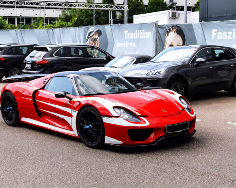 Roter Porsche-Supersportwagen steht vor mehreren schwarzen Porsche-SUVs auf einem Parkplatz.