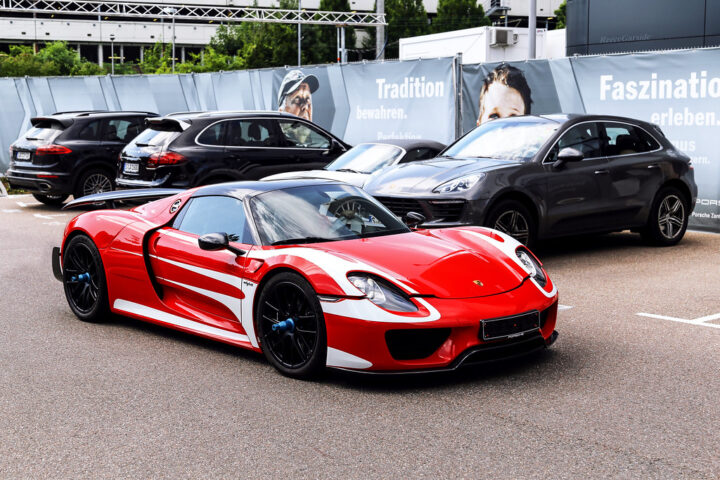 Roter Porsche-Supersportwagen steht vor mehreren schwarzen Porsche-SUVs auf einem Parkplatz.