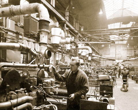 Arbeiter in einer Industriehalle zwischen Maschinen und Rohrleitungen.