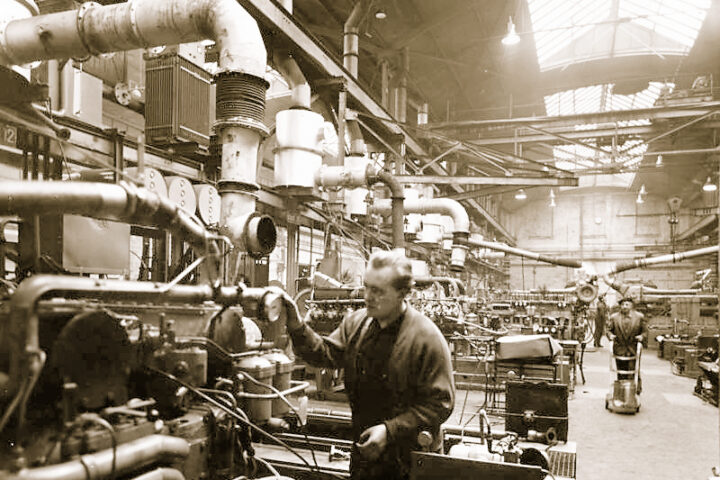 Arbeiter in einer Industriehalle zwischen Maschinen und Rohrleitungen.