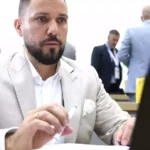 Tobias Rausch sitzt in heller Kleidung an einem Tisch im Parlament und hält einen Stift in der Hand.