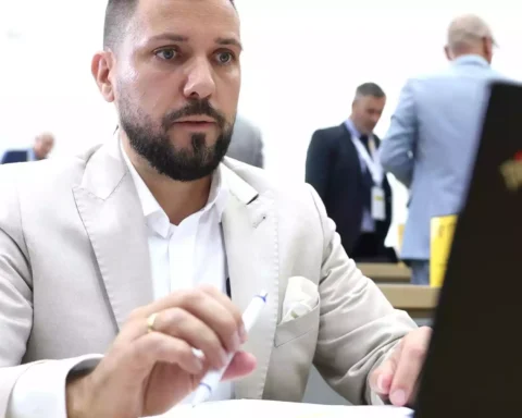 Tobias Rausch sitzt in heller Kleidung an einem Tisch im Parlament und hält einen Stift in der Hand.
