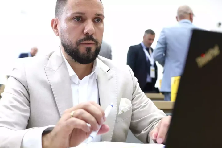 Tobias Rausch sitzt in heller Kleidung an einem Tisch im Parlament und hält einen Stift in der Hand.