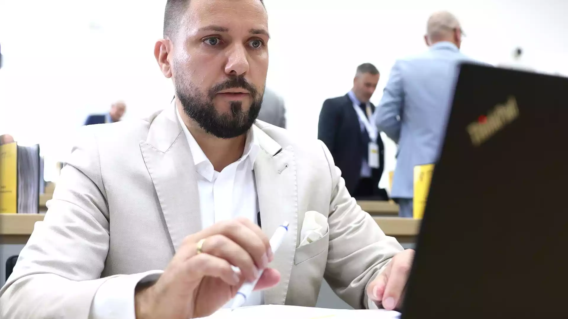 Tobias Rausch sitzt in heller Kleidung an einem Tisch im Parlament und hält einen Stift in der Hand.