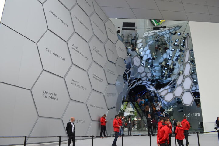 Besucher vor einem Audi-Exponat mit hexagonalem Wanddesign und Spiegelkonstruktion auf einer Automesse.