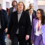 Mehrere Personen gehen in einem Innenraum, zwei Frauen im Vordergrund tragen Businesskleidung und Besucherausweise.