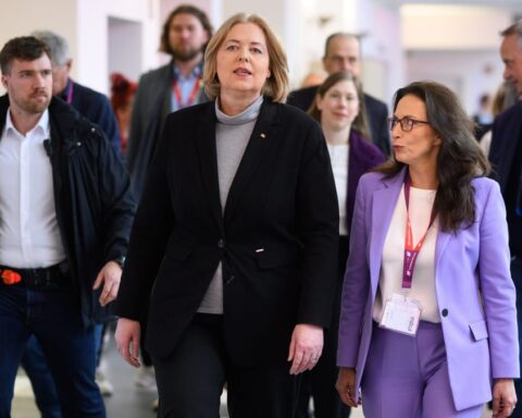 Mehrere Personen gehen in einem Innenraum, zwei Frauen im Vordergrund tragen Businesskleidung und Besucherausweise.