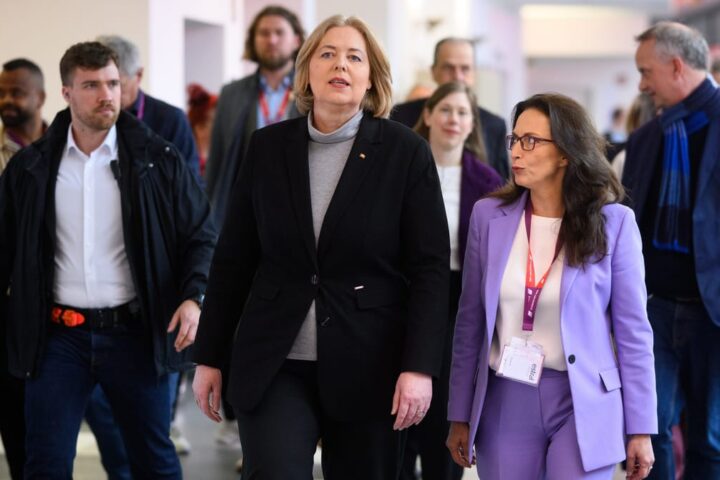 Mehrere Personen gehen in einem Innenraum, zwei Frauen im Vordergrund tragen Businesskleidung und Besucherausweise.
