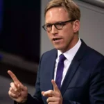 Mann im Anzug mit Brille spricht gestikulierend vor grauem Hintergrund.