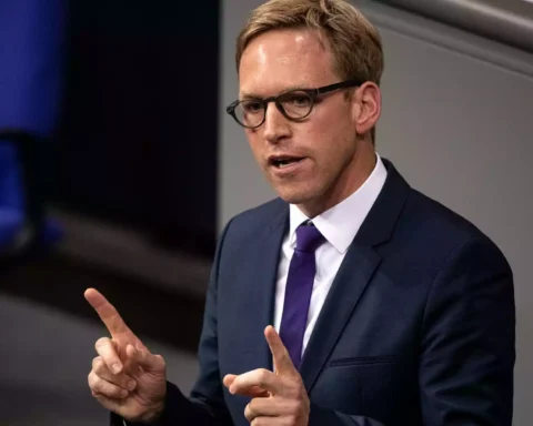 Mann im Anzug mit Brille spricht gestikulierend vor grauem Hintergrund.