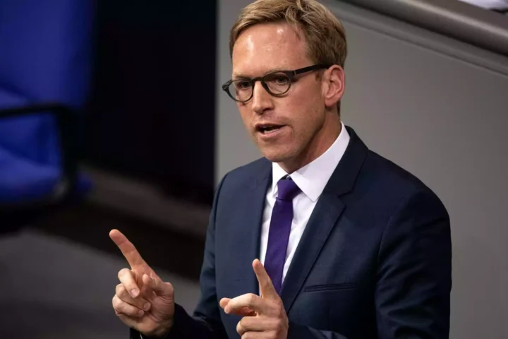 Mann im Anzug mit Brille spricht gestikulierend vor grauem Hintergrund.