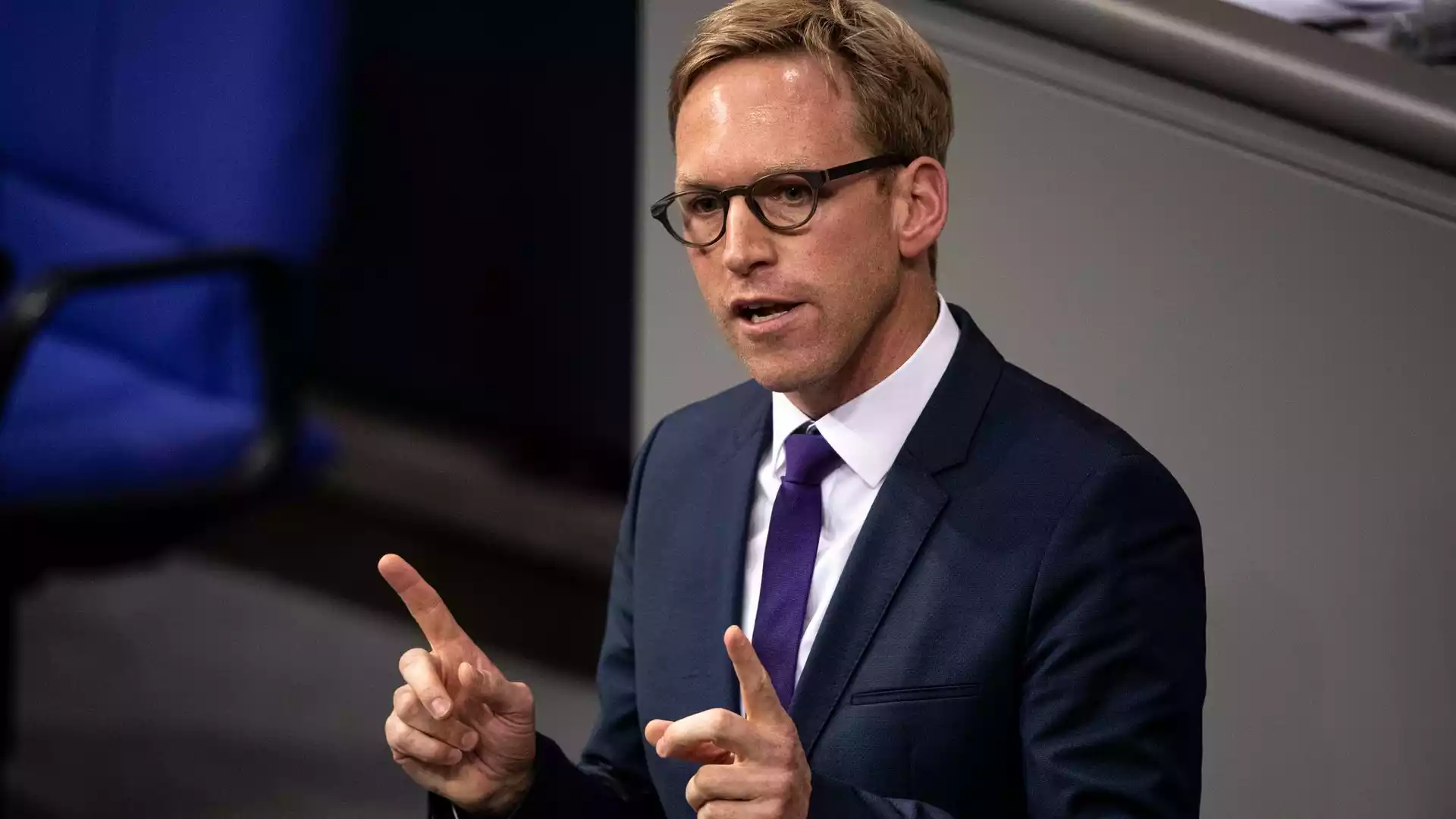 Mann im Anzug mit Brille spricht gestikulierend vor grauem Hintergrund.