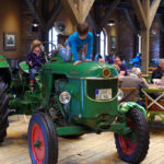 Grüner Deutz-Traktor in einem Innenraum mit mehreren Sitzenden im Hintergrund.