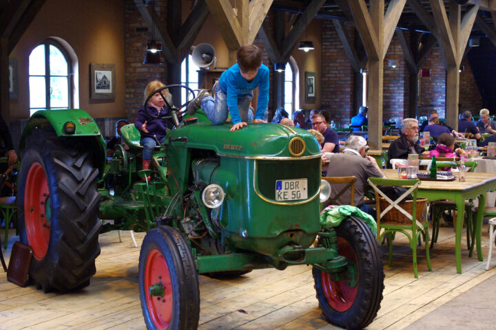 Grüner Deutz-Traktor in einem Innenraum mit mehreren Sitzenden im Hintergrund.