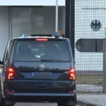 Ein dunkler VW fährt vor einem gesicherten Tor mit Bundesadler an einem Behördengebäude vor.