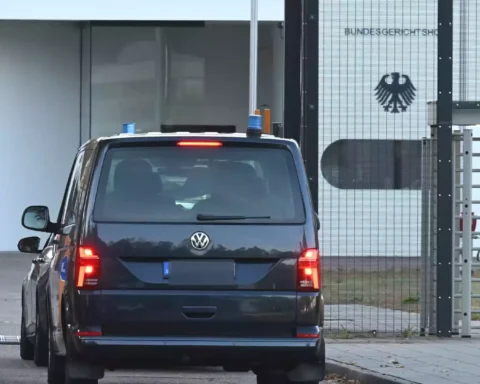 Ein dunkler VW fährt vor einem gesicherten Tor mit Bundesadler an einem Behördengebäude vor.
