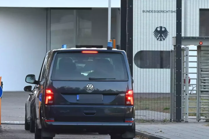 Ein dunkler VW fährt vor einem gesicherten Tor mit Bundesadler an einem Behördengebäude vor.