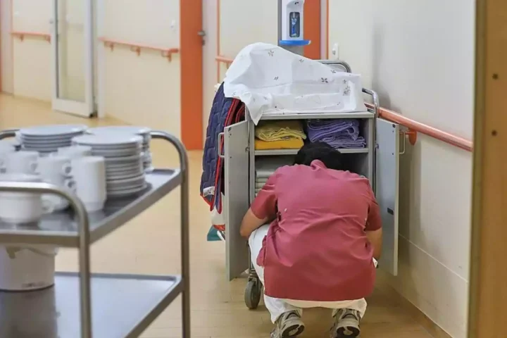 Eine Pflegekraft kniet neben einem Wäsche- oder Materialwagen in einem Klinikflur.