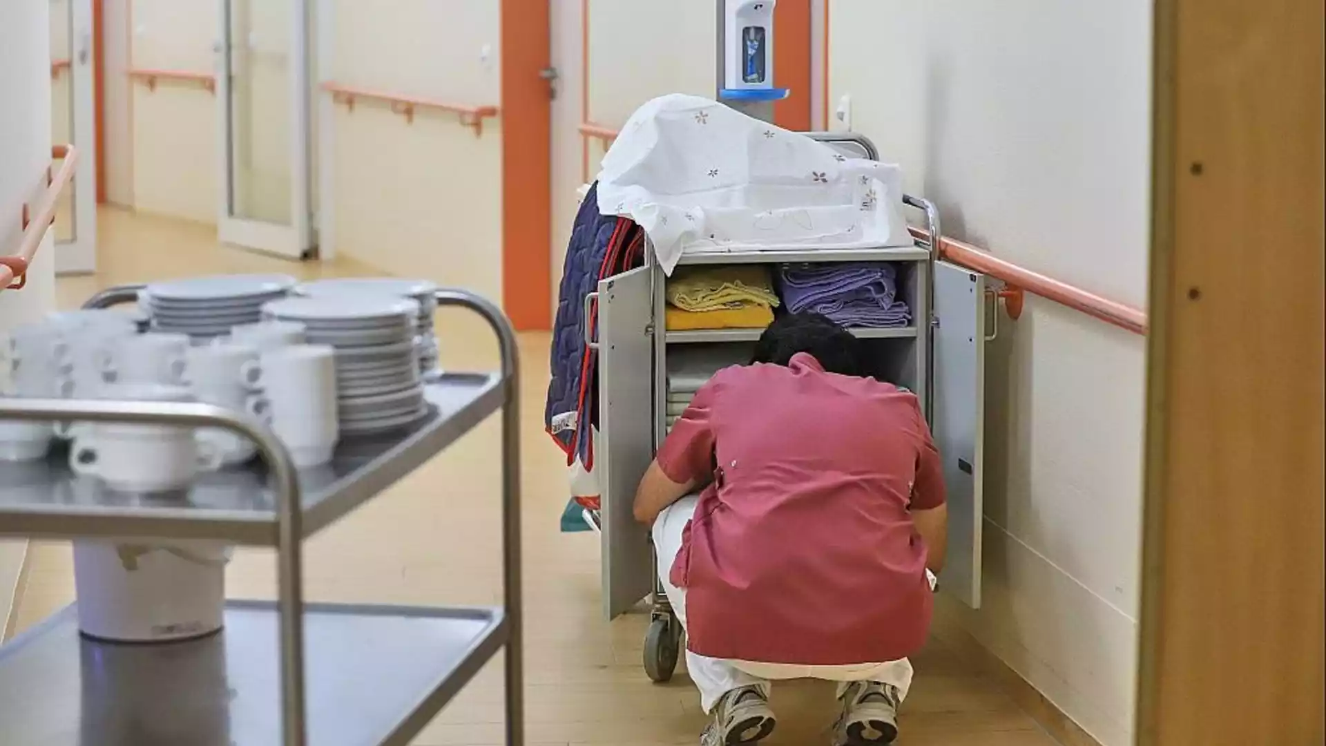 Linnemann sieht Pflegestrukturkommission vor dem Abschluss 1 Eine Pflegekraft kniet neben einem Wäsche- oder Materialwagen in einem Klinikflur.
