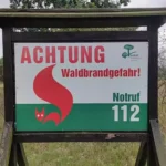 Warnschild mit der Aufschrift „Achtung Waldbrandgefahr“ und Notruf 112 an einem Waldrand.