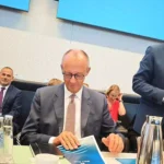 Mehrere Politiker sitzen in einem Sitzungssaal an einem Tisch; ein Mann im Anzug spricht am Mikrofon.