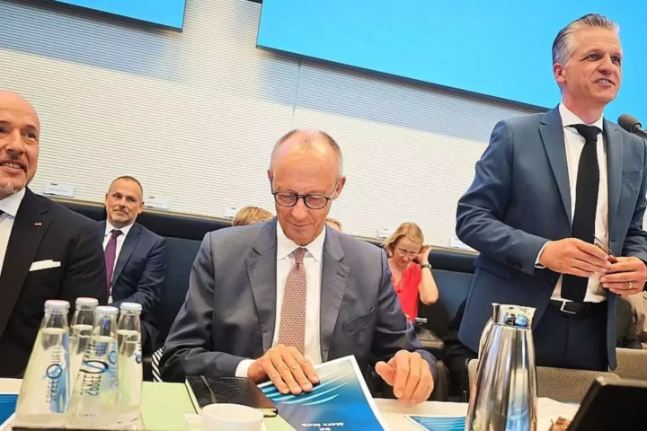 Mehrere Politiker sitzen in einem Sitzungssaal an einem Tisch; ein Mann im Anzug spricht am Mikrofon.