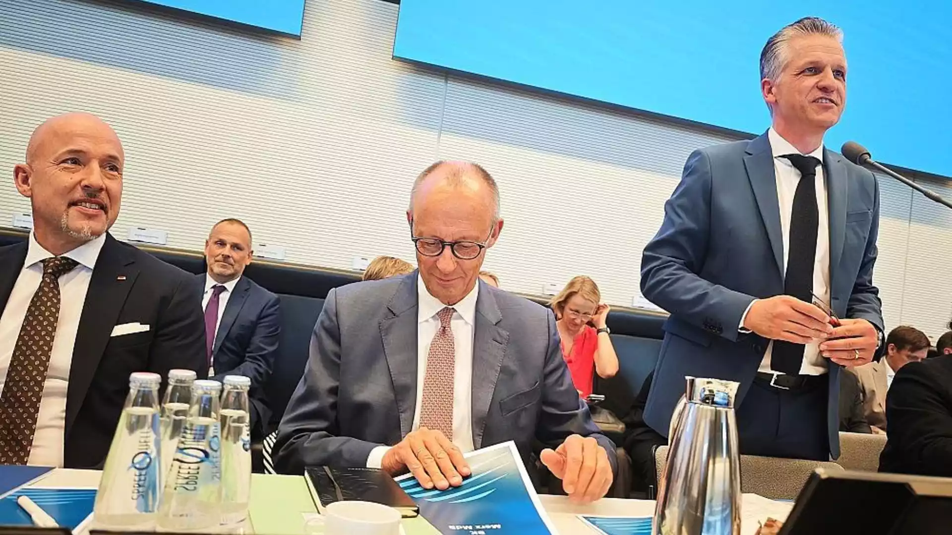 Mehrere Politiker sitzen in einem Sitzungssaal an einem Tisch; ein Mann im Anzug spricht am Mikrofon.
