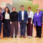 Sieben Personen stehen auf einer Bühne vor einer bunten Wanddekoration.