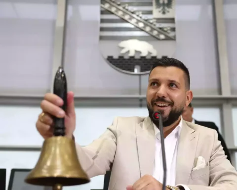 Mann in hellem Anzug läutet in einem Parlamentssaal eine Glocke am Rednerpult.