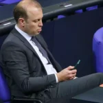 Mann im Anzug sitzt in einem Parlamentssaal und schaut auf ein Smartphone.