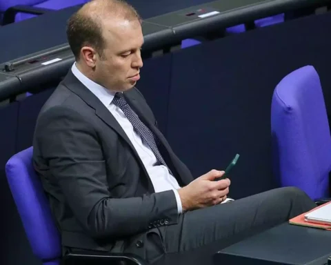 Mann im Anzug sitzt in einem Parlamentssaal und schaut auf ein Smartphone.