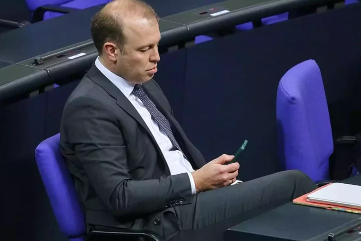 Mann im Anzug sitzt in einem Parlamentssaal und schaut auf ein Smartphone.