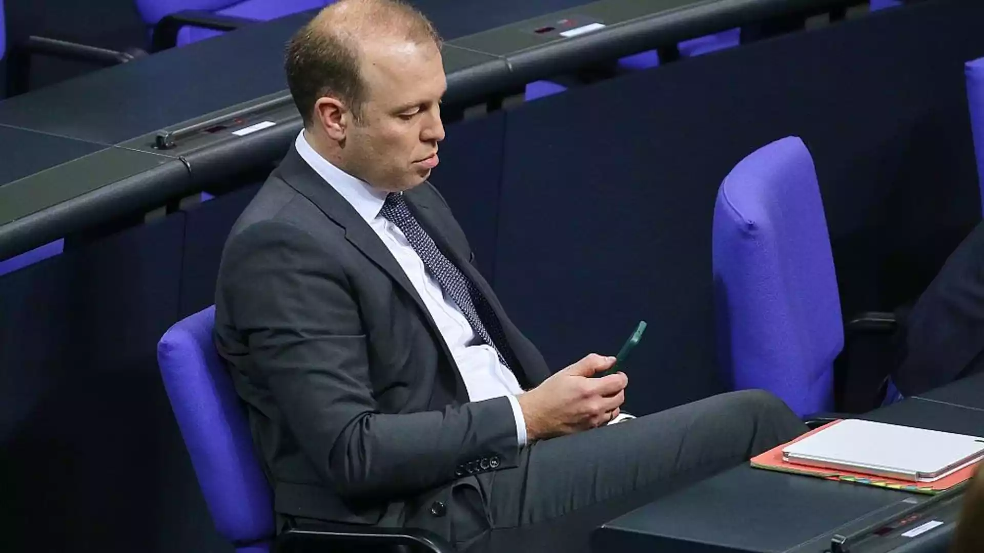 Mann im Anzug sitzt in einem Parlamentssaal und schaut auf ein Smartphone.