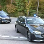 Zwei schwarze Fahrzeuge mit Blaulicht fahren auf einer Straße durch ein bewaldetes Gebiet.