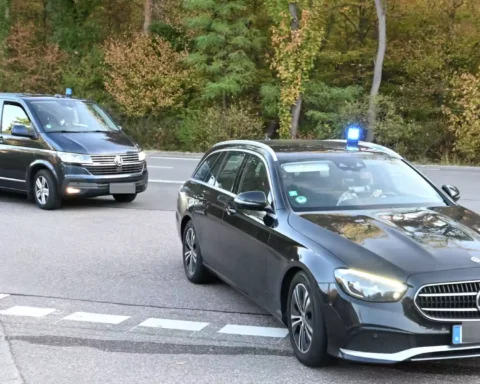 Zwei schwarze Fahrzeuge mit Blaulicht fahren auf einer Straße durch ein bewaldetes Gebiet.