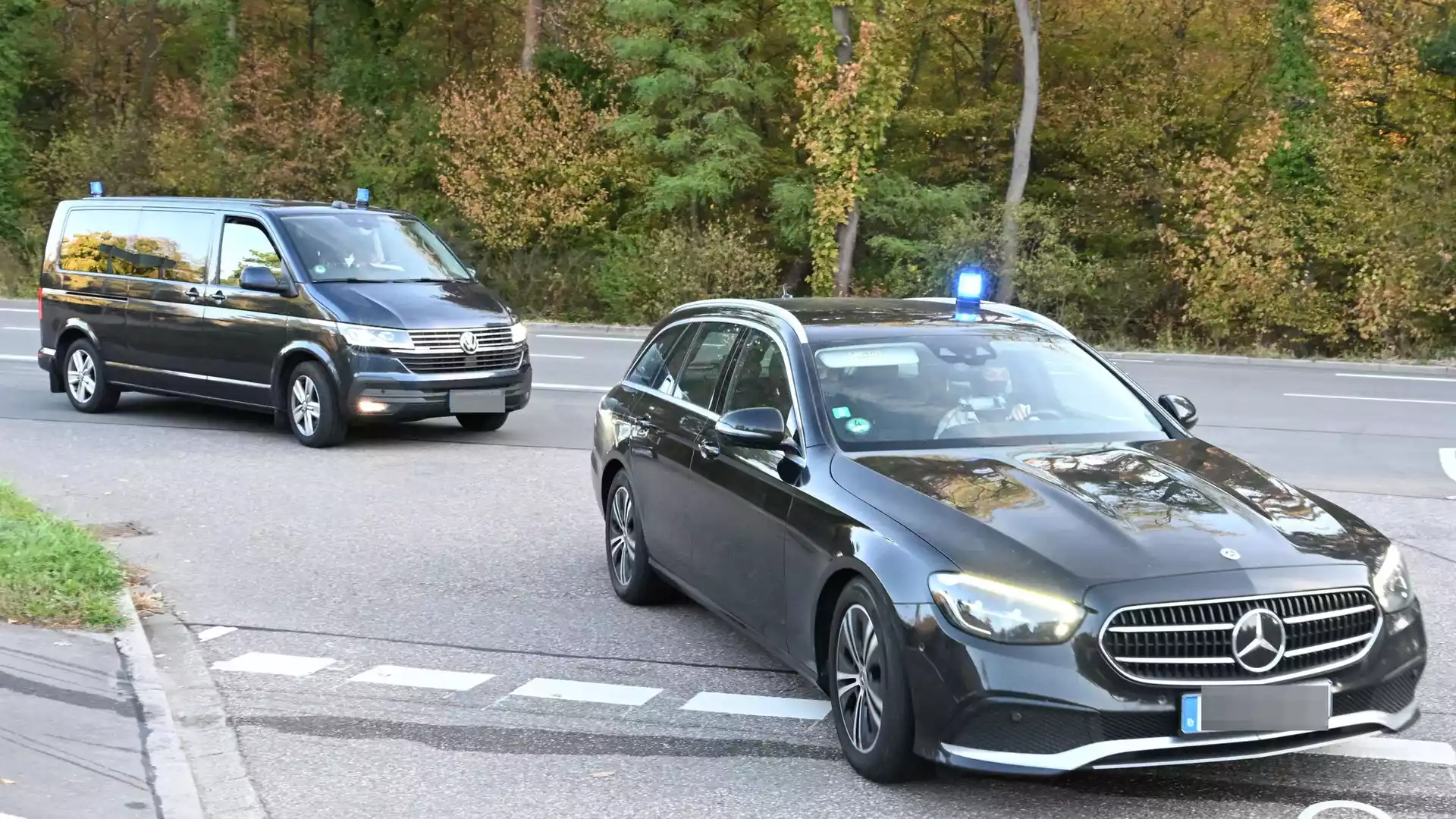Zwei schwarze Fahrzeuge mit Blaulicht fahren auf einer Straße durch ein bewaldetes Gebiet.