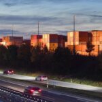 Containerterminal neben einer Autobahn bei Abendlicht, mit gestapelten Containern und vorbeifahrenden Autos.