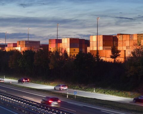 Containerterminal neben einer Autobahn bei Abendlicht, mit gestapelten Containern und vorbeifahrenden Autos.