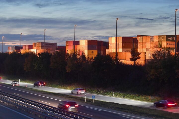 Containerterminal neben einer Autobahn bei Abendlicht, mit gestapelten Containern und vorbeifahrenden Autos.