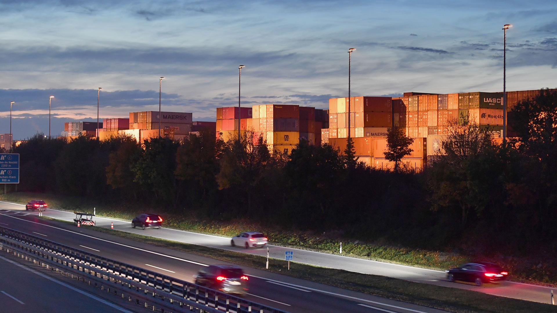Containerterminal neben einer Autobahn bei Abendlicht, mit gestapelten Containern und vorbeifahrenden Autos.