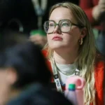 Eine Frau mit großer Brille sitzt aufmerksam in einer Menschenmenge bei einer Veranstaltung.